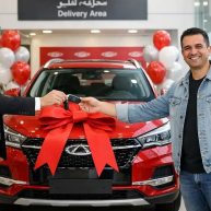تصویری از لحظه تحویل خودرو مشتری کنار ماشین قرمز با روبان در حال دریافت کلید در نمایشگاه